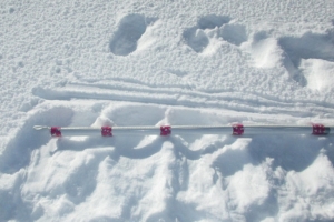 Close-up of a white SIMBA thermistor chain laid horizontally across a silver support pole on snow. Red spacing bands secure the chain at regular intervals, with footprints, equipment tracks and disturbed snow visible around the installation area. Close-up of a white SIMBA thermistor chain laid horizontally across a silver support pole on snow. Red spacing bands secure the chain at regular intervals, with footprints, equipment tracks and disturbed snow visible around the installation area.
