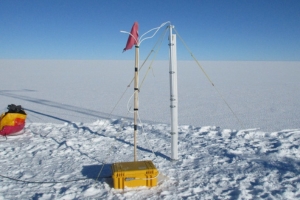 SIMBA monitoring equipment installed on the snow-covered Greenland Ice Sheet beneath a clear blue sky. A vertical white thermistor chain is secured beside a guyed support mast and connected by cables to a yellow protective equipment case on the snow. A red marker flag identifies the installation. SIMBA monitoring equipment installed on the snow-covered Greenland Ice Sheet beneath a clear blue sky. A vertical white thermistor chain is secured beside a guyed support mast and connected by cables to a yellow protective equipment case on the snow. A red marker flag identifies the installation.