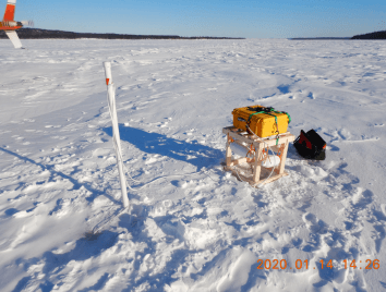 SIMBA monitoring unit installed on a raised platform on the snow-covered Churchill River in Canada, with a marker pole and field equipment nearby.