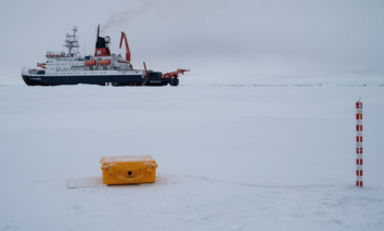Research icebreaker ship anchored on a frozen sea with a yellow SIMBA snow-and ice monitoring device and a red-and-white measurement pole in the snow.