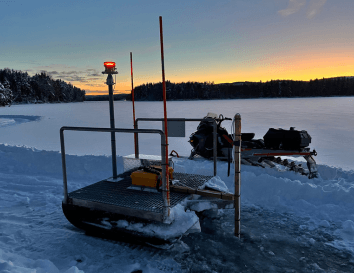 SIMBA snow-and ice monitoring device and a snowmobile on a frozen lake at sunset with a snow-covered forest in the background. SIMBA snow-and ice monitoring device and a snowmobile on a frozen lake at sunset with a snow-covered forest in the background.
