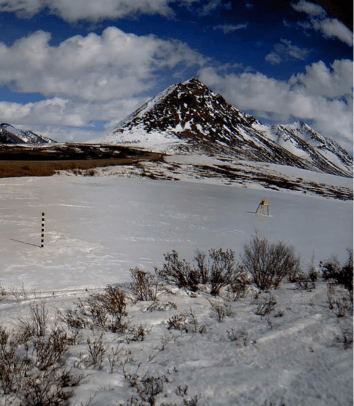 Snow-covered mountain under a partly cloudy blue sky with sparse vegetation and a yellow SIMBA snow- and ice monitoring device with measuring poles in the foreground. Snow-covered mountain under a partly cloudy blue sky with sparse vegetation and a yellow SIMBA snow- and ice monitoring device with measuring poles in the foreground.