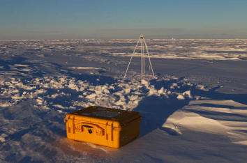 Yellow SIMBA snow-and ice monitoring device on icy ground in a vast, snowy landscape with a tripod structure in the background under clear sky. Yellow SIMBA snow-and ice monitoring device on icy ground in a vast, snowy landscape with a tripod structure in the background under clear sky.