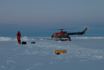Helicopter landed on snowy ice with two people in orange suits nearby and a yellow SIMBA snow- and ice monitoring device on the ground under a clear sky. Helicopter landed on snowy ice with two people in orange suits nearby and a yellow SIMBA snow- and ice monitoring device on the ground under a clear sky.