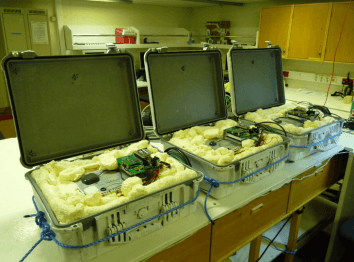 Three open, foam-lined cases on a bench, each containing electronic components and circuit boards in a lab setting.