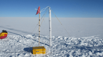 SIMBA snow- and ice monitoring device with a wind sock and instruments set up on a vast, snowy, and icy landscape under a clear sky.