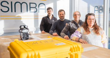 Four people a table with a yellow SIMBA case and a pressure gauge device, in a bright room with large windows.